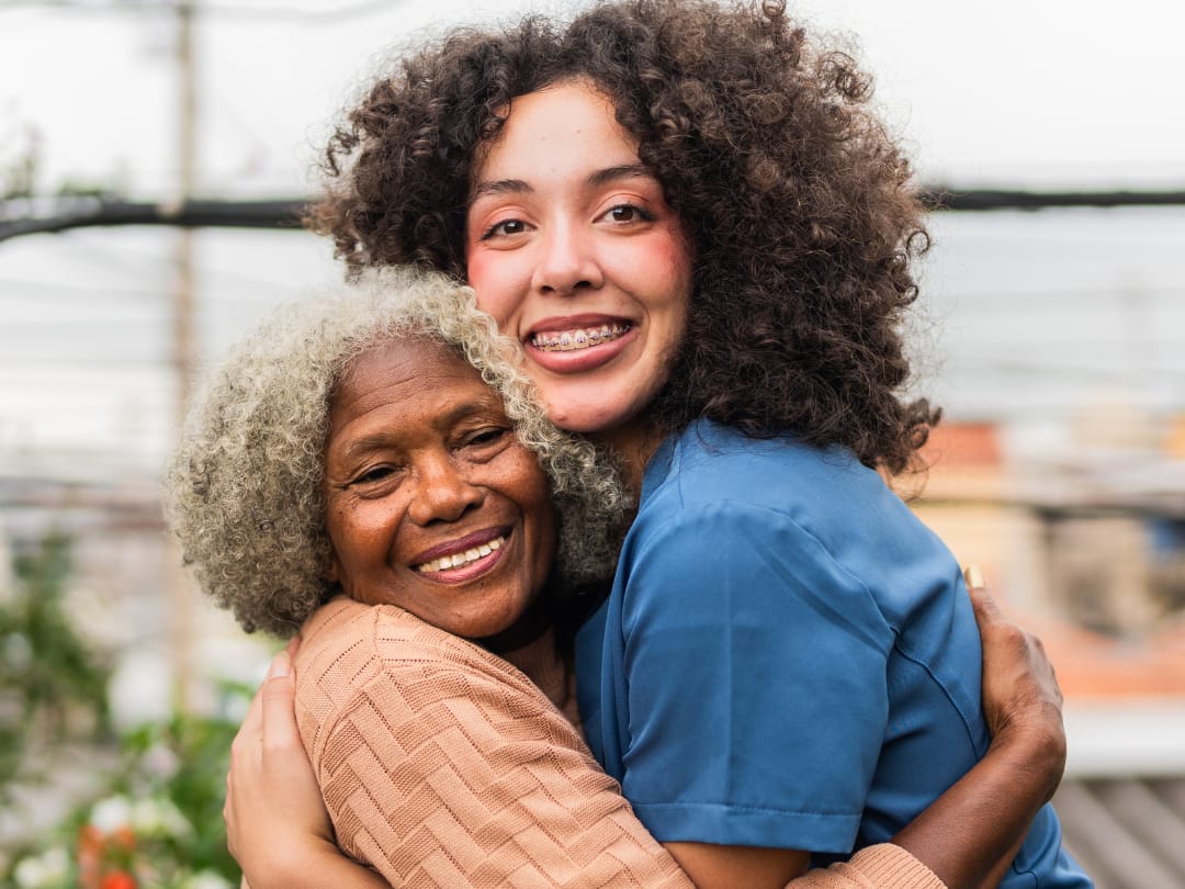 An older woman and a young woman in blue scrubs hug and smile outdoors.