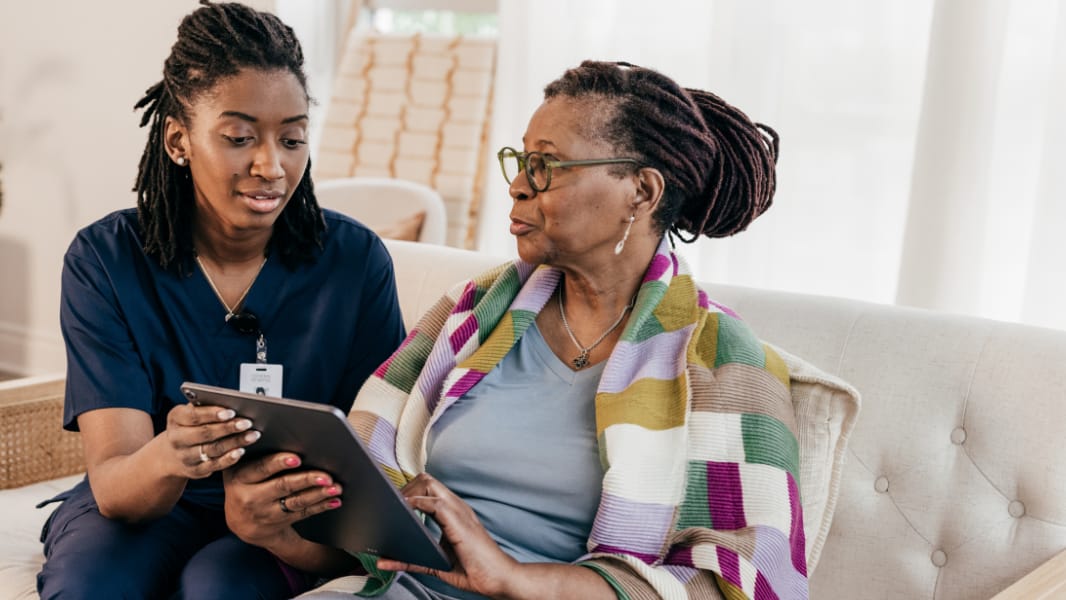 A nurse in navy scrubs shows an older woman something on a tablet as they sit together on a sofa.